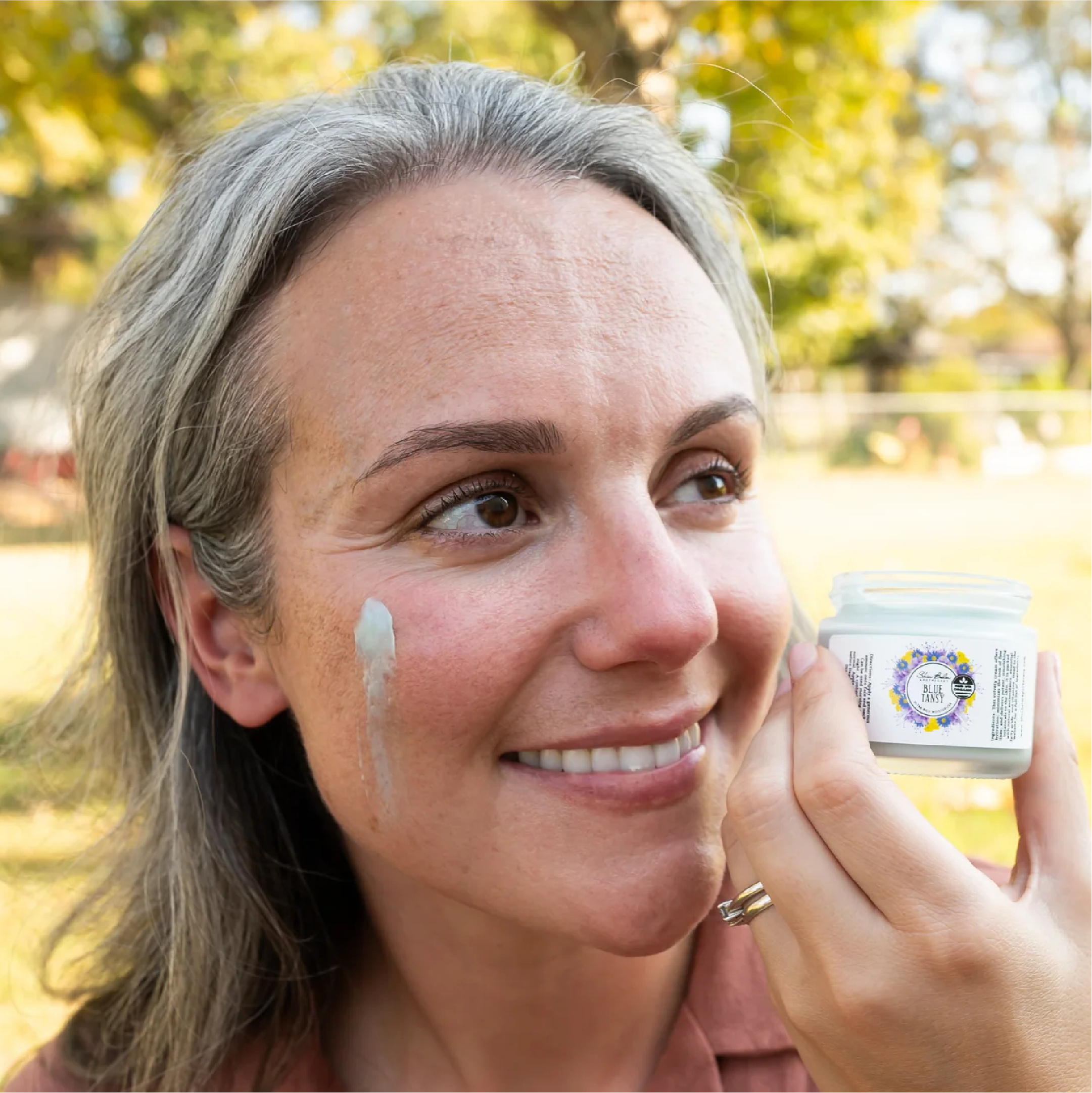 A smiling woman holds an open jar of Blue Tansy Ultra Rich Moisturizer outdoors with the product on her cheek.