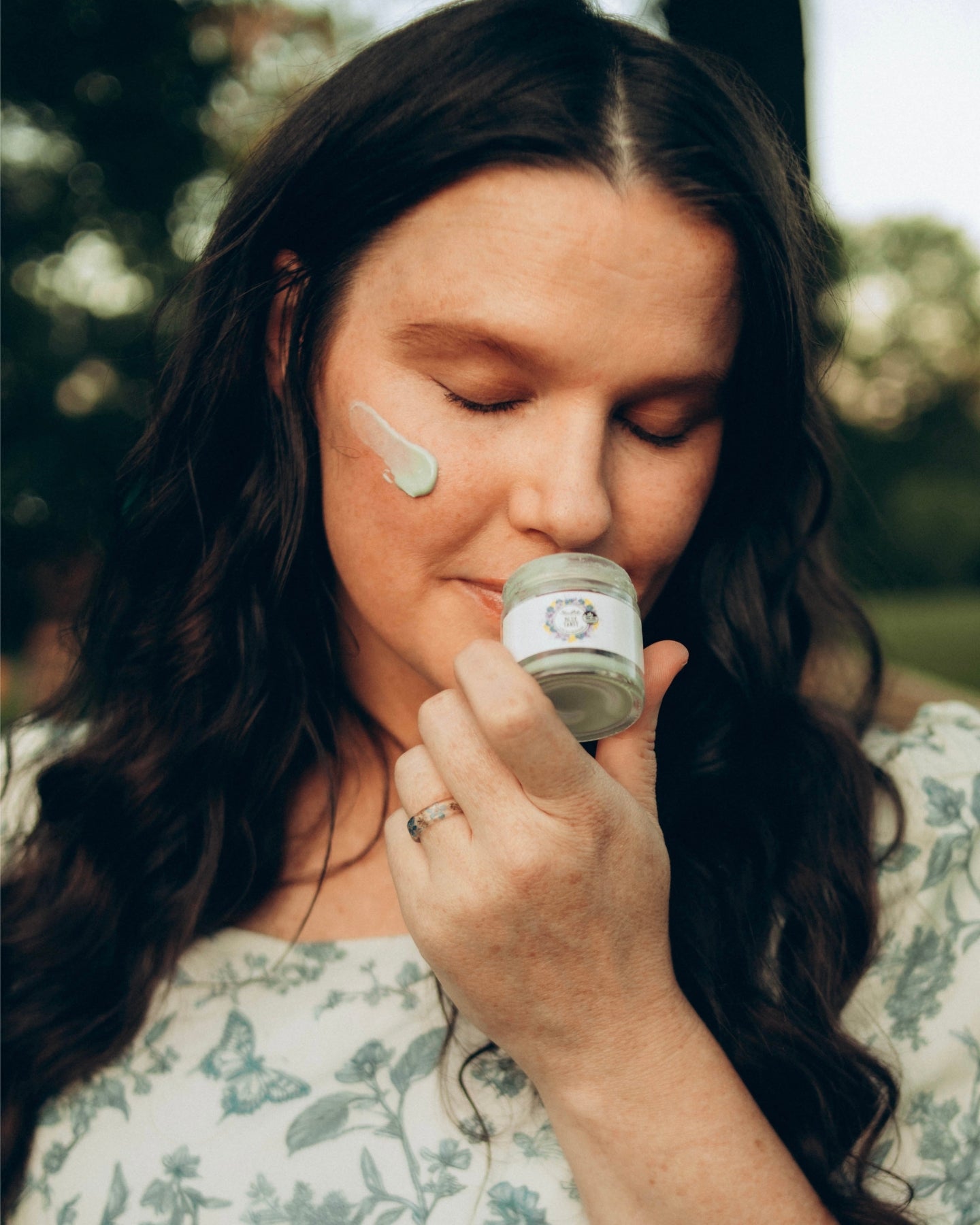 A woman closes her eyes and smells an open jar of Blue Tansy Ultra Rich Moisturizer with a daub of the product on her cheek.