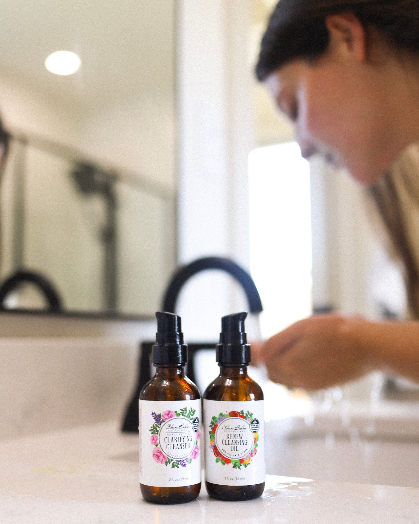 Facial Cleansing Duo resting on a bathroom sink focused in the foreground with a woman washing her face blurred in the background.